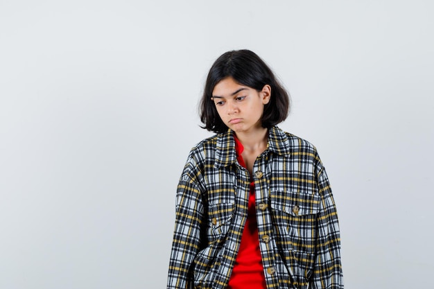 Jeune fille debout, regardant vers le bas et se présentant à la caméra en chemise à carreaux et t-shirt rouge et l'air songeur. vue de face.