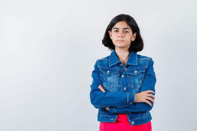 Jeune fille debout tout droit et se présentant à la caméra en t-shirt rouge et veste en jean et l'air sérieux, vue de face.