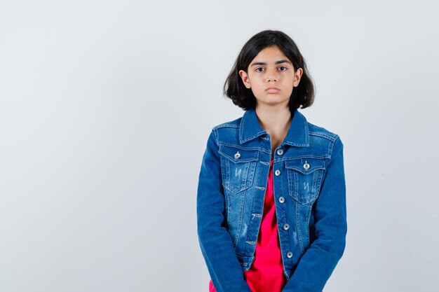 Jeune fille debout tout droit et se présentant à la caméra en t-shirt rouge et veste en jean et l'air sérieux.