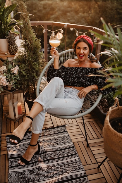 Photo gratuite jeune fille élégante aux cheveux ondulés et bandeau lumineux en haut noir, jeans clairs et talons noirs assis et tenant un cocktail sur une terrasse moderne..