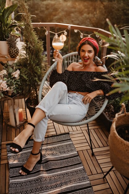 Jeune fille élégante aux cheveux ondulés et bandeau lumineux en haut noir, jeans clairs et talons noirs assis et tenant un cocktail sur une terrasse moderne..