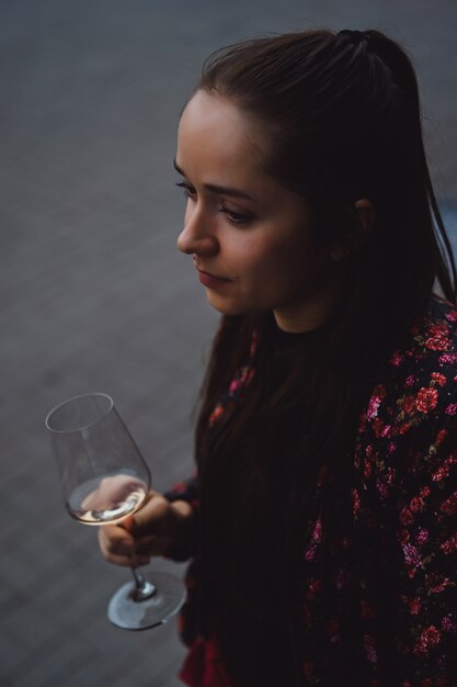 Une jeune fille élégante boit du vin dans un café de rue sur une terrasse d&#39;été. Une fille aux cheveux longs apprécie un verre de vin sur une soirée d&#39;été. Portrait. fermer