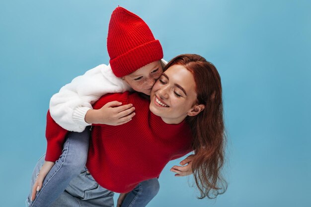 Jeune fille élégante avec des taches de rousseur en tenue blanche et une tasse rouge embrassant sa soeur souriante en pull brillant sur fond bleu