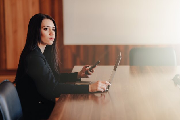 Jeune fille émotive attrayante dans les vêtements de style d'affaires assis à un bureau sur un ordinateur portable et un téléphone dans le bureau ou l'auditorium