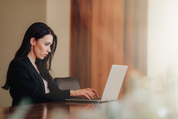 Jeune fille émotive attrayante dans les vêtements de style business assis à un bureau sur un ordinateur portable et un téléphone dans le bureau ou l&#39;auditorium