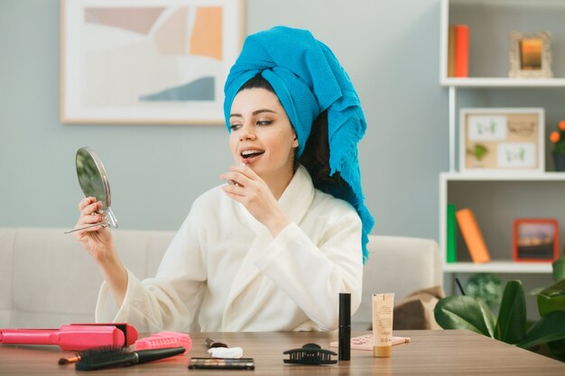 Jeune fille enveloppée de cheveux dans une serviette appliquant du rouge à lèvres tenant et regardant un miroir assis à table avec des outils de maquillage dans le salon