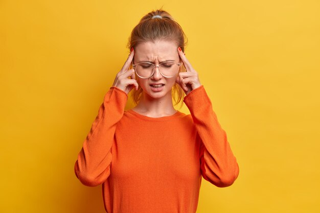 Jeune fille épuisée touche les tempes, souffre de maux de tête insupportables, fronce les sourcils, porte de grandes lunettes optiques, cavalier orange, se tient à l'intérieur