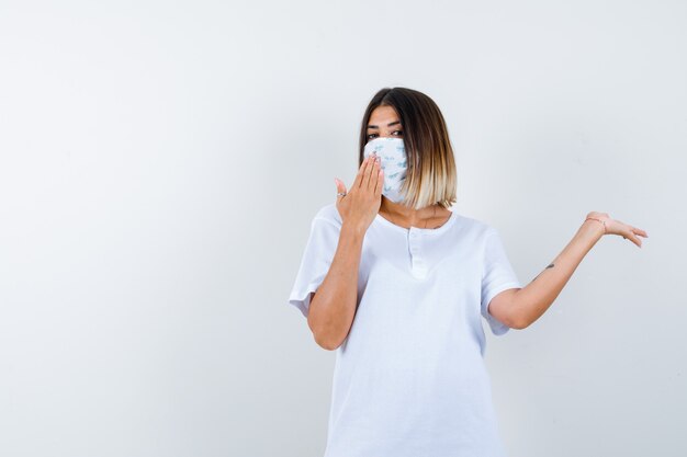 Jeune fille étalant la paume de côté, montrant un panneau d'arrêt en t-shirt blanc et masque et à la vue de face, confiant.