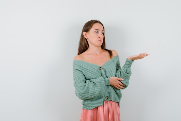 Jeune fille étalant la paume de côté, tenant la main sur le coude en maille, jupe et à l'ennui. vue de face.