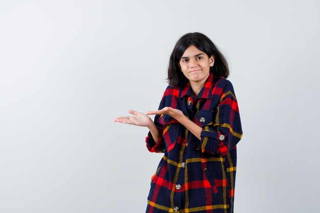 Jeune fille étirant la main comme présentant et tenant quelque chose en chemise à carreaux et l'air heureux. vue de face.