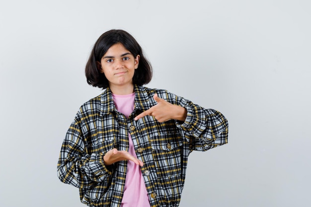 Jeune Fille étirant Une Main Comme Tenant Quelque Chose Et Pointant Vers Elle Avec L'index En Chemise à Carreaux Et T-shirt Rose Et L'air Mignonne, Vue De Face.