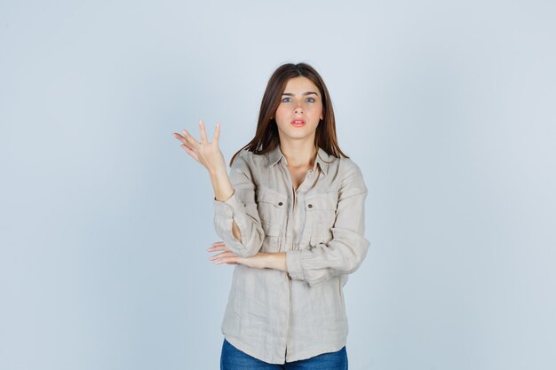 Jeune fille étirant la main de manière interrogative en chemise beige, jeans et l'air perplexe, vue de face.