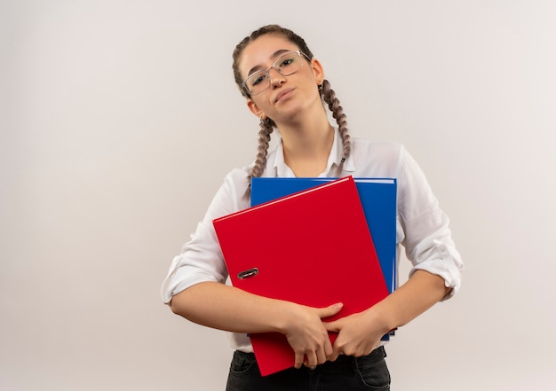 Jeune fille étudiante dans des verres avec des nattes en chemise blanche dossiers hlding à l'avant mécontent debout sur mur blanc