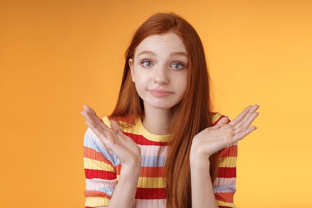 Une jeune fille européenne idiote rousse sans aucune idée des années 20, haussant les mains, se propage sur le côté avec un sourire narquois désolé ne peut pas répondre debout, inconscient, confus, perplexe, donne une réponse, fond orange.