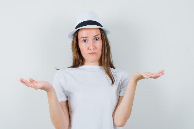 Jeune fille faisant le geste des échelles en t-shirt blanc, chapeau et à la vue indécise, de face.