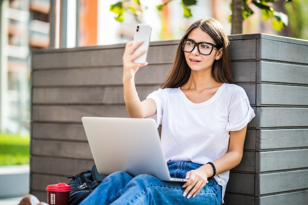 Jeune fille faisant selfie sur téléphone mobile ou appel vidéo. Femme assise sur un banc travaillant sur un ordinateur portable pc moderne dans le parc de la ville dans la rue à l'extérieur sur la nature. Concept d'entreprise indépendant.