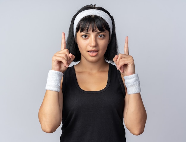 Jeune fille fitness portant un bandeau regardant la caméra heureuse et surprise montrant les index debout sur blanc