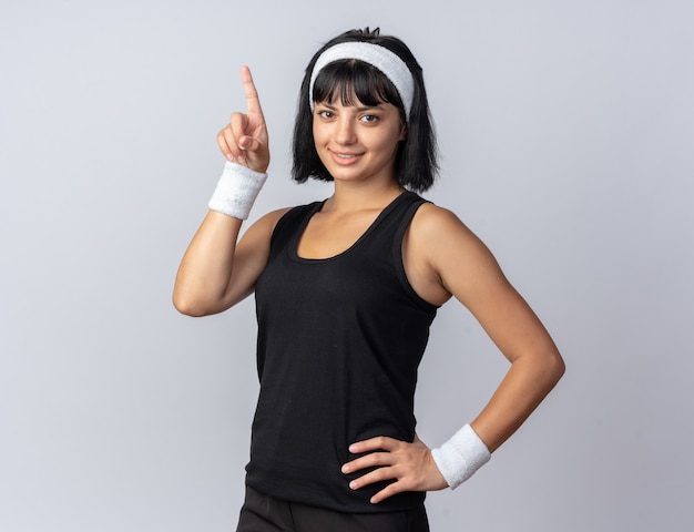 Jeune fille fitness portant un bandeau regardant la caméra souriante confiante pointant avec l'index vers le haut debout sur blanc