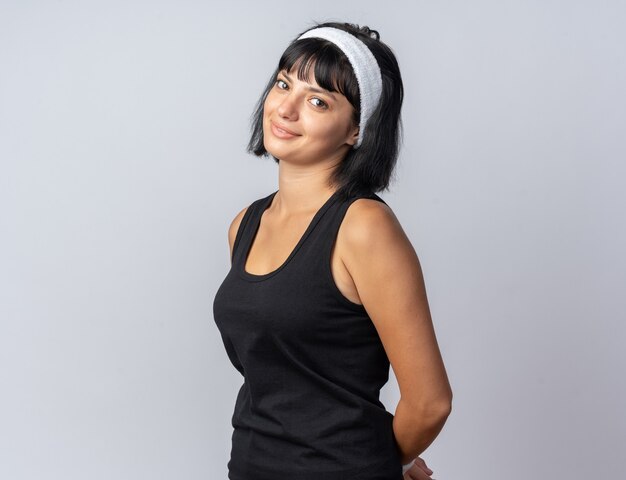 Jeune fille fitness portant un bandeau regardant la caméra avec un sourire timide sur le visage debout sur fond blanc