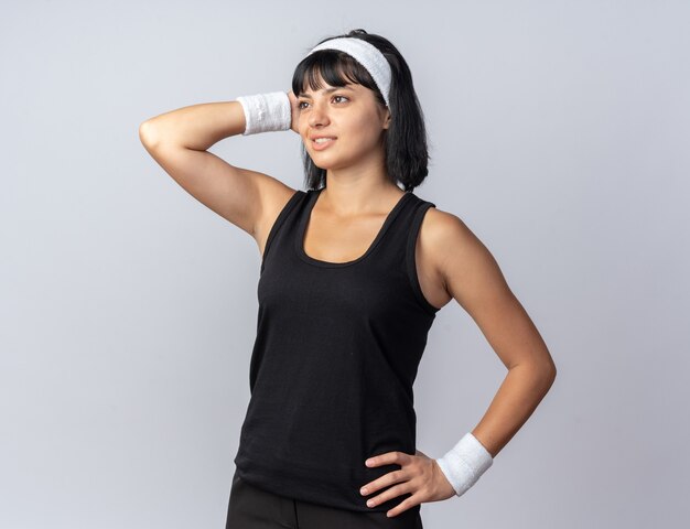 Jeune fille fitness portant un bandeau regardant de côté souriant tenant la main sur sa tête pensant positivement debout sur blanc