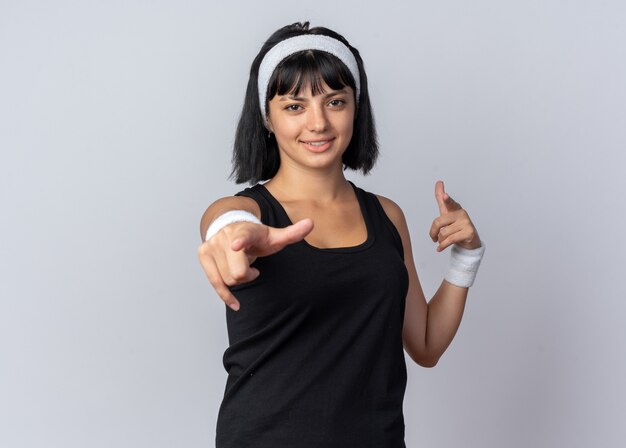 Jeune fille fitness portant un bandeau souriant pointant gaiement avec l'index à la caméra debout sur fond blanc