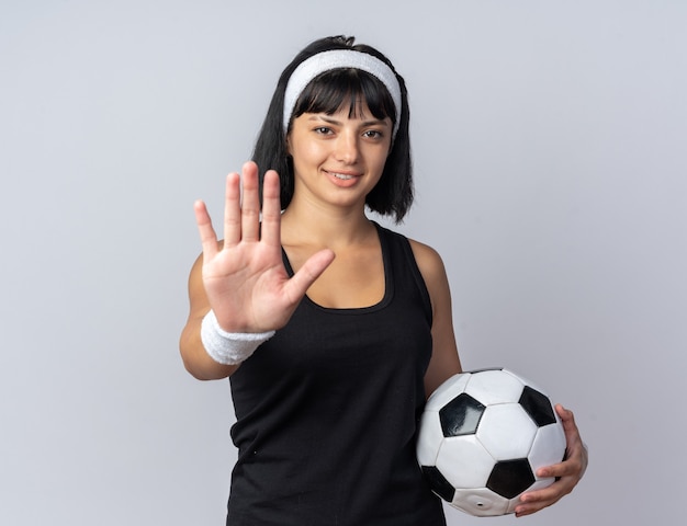 Jeune fille fitness portant un bandeau tenant un ballon de football regardant la caméra souriant faisant un geste d'arrêt avec la main debout sur blanc