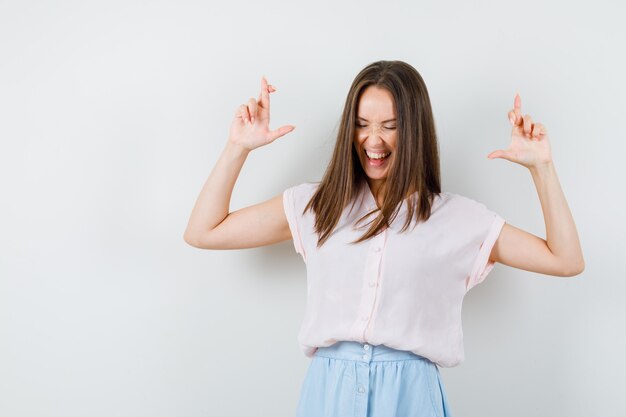 Jeune fille en gardant les doigts croisés en t-shirt, jupe et regardant heureux, vue de face.