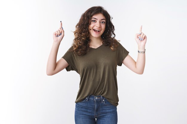 Une jeune fille géorgienne créative excitée et excitée aux cheveux bouclés souriante vous encourage à essayer le produit pointant l'index vers le haut pour promouvoir une publicité cool, fond blanc