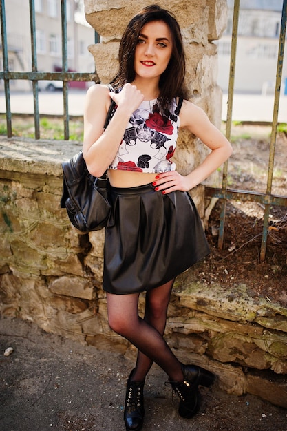 Jeune fille goth sur jupe en cuir noir et chaussures punk à talons hauts avec sac à dos posé contre une clôture de fer