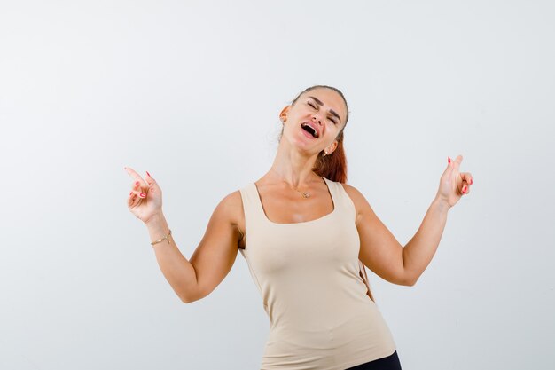 Jeune fille en haut beige, pantalon noir pointant vers le haut avec l'index et l'air heureux, vue de face.