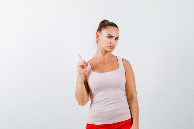 Jeune fille en haut beige et pantalon rouge levant l'index en geste eureka et à la vue sensible, de face.