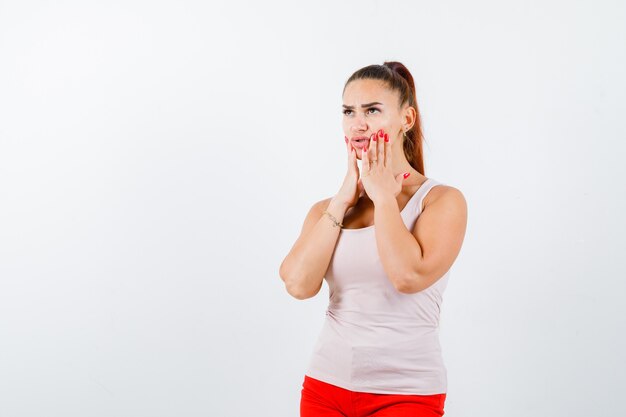 Jeune fille en haut beige et pantalon rouge tenant les mains sur les joues et à la vue de face, surpris.