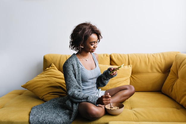 Jeune fille heureuse, petit-déjeuner sain, gruau, granola, écouter de la musique dans des écouteurs sur un canapé jaune à la maison. Elle a les cheveux courts et bouclés, porte un long cardigan gris, un short.