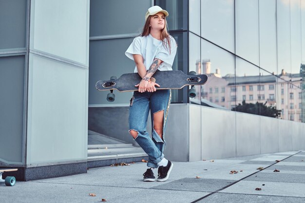 Jeune fille hipster en casquette vêtue d'une chemise blanche et d'un jean déchiré tient une planche à roulettes tout en posant près d'un gratte-ciel.