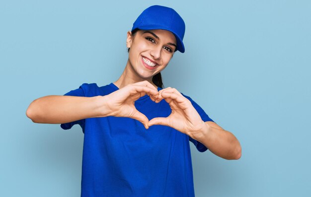 Jeune fille hispanique portant l'uniforme de courrier de livraison souriant amoureux faisant la forme de symbole de coeur avec les mains. notion romantique.