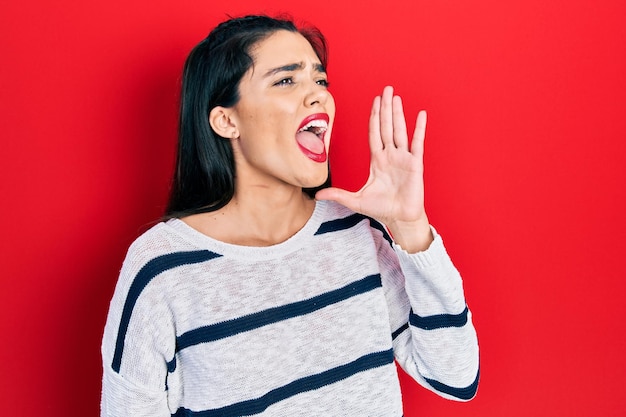 Jeune fille hispanique portant des vêtements décontractés criant et criant fort à côté avec le concept de communication main sur bouche