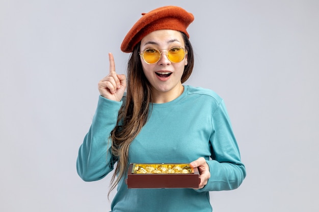 Jeune fille impressionnée le jour de la Saint-Valentin portant un chapeau avec des lunettes tenant une boîte de bonbons pointe vers le haut isolé sur fond blanc