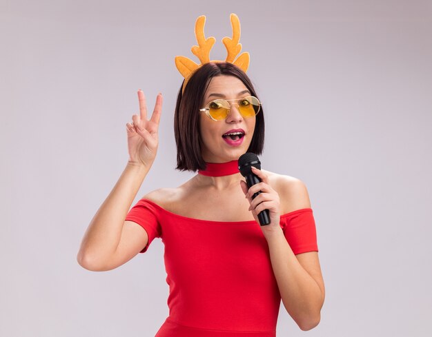 Jeune fille impressionnée portant un bandeau en bois de renne et des lunettes parlant dans un microphone regardant la caméra faisant un signe de paix isolé sur fond blanc