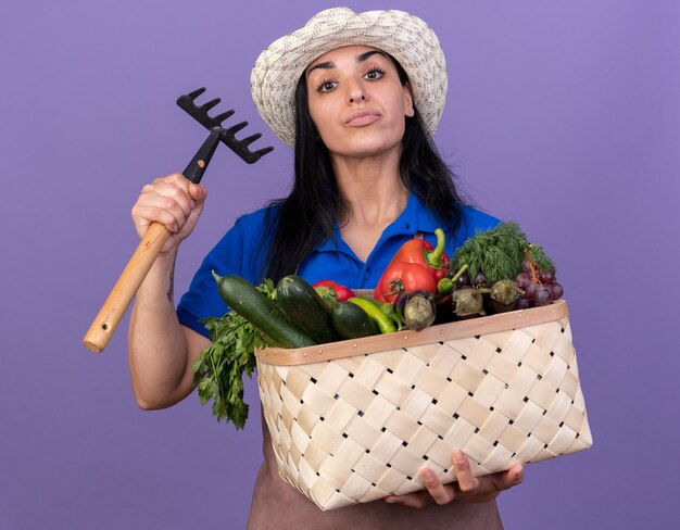 Jeune fille de jardinier caucasienne confiante en uniforme et chapeau tenant un panier de légumes et un râteau