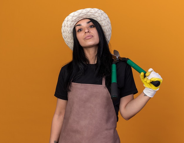Jeune fille de jardinier confiante portant un uniforme et un chapeau avec des gants de jardinier tenant des cisailles à haie sur l'épaule regardant à l'avant isolé sur un mur orange avec espace de copie