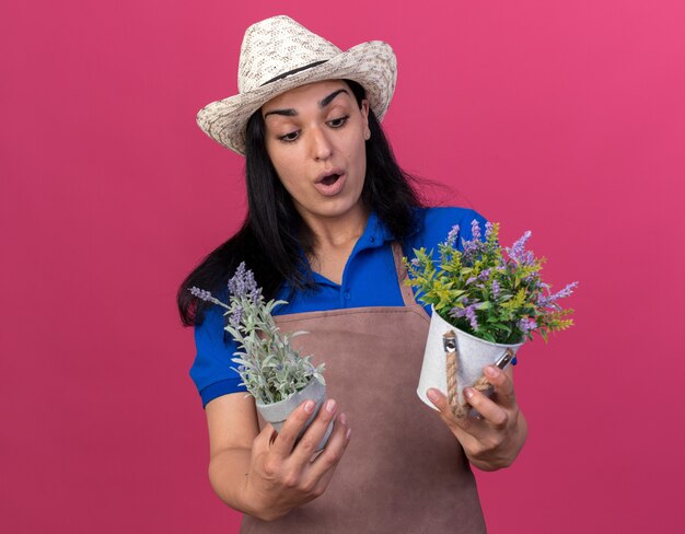 Jeune fille de jardinier surprise en uniforme et chapeau tenant et regardant des pots de fleurs isolés sur un mur rose