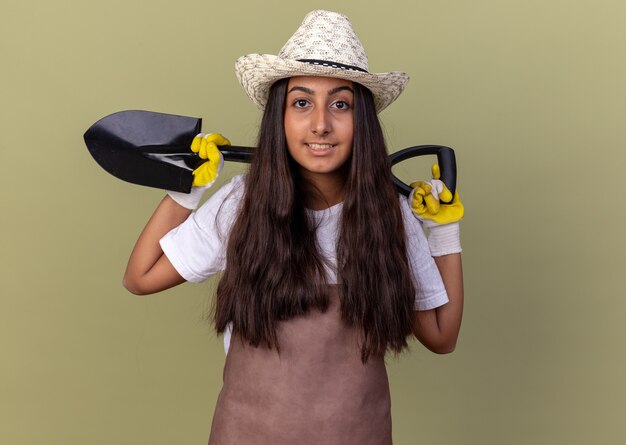 Jeune fille de jardinier en tablier et chapeau d'été portant des gants de travail tenant une pelle souriant debout confiant sur mur vert