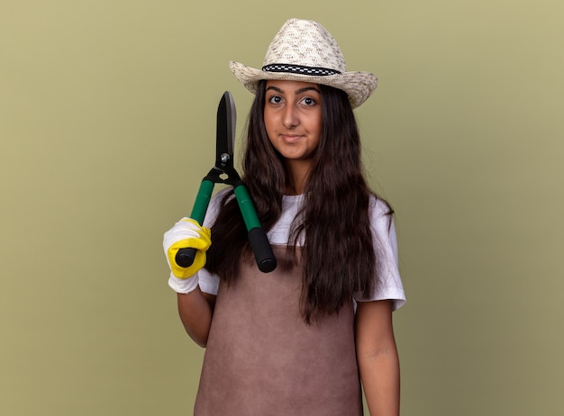 Jeune fille de jardinier en tablier et chapeau d'été portant des gants de travail tenant une tondeuse à haies smiling confiant debout sur mur vert