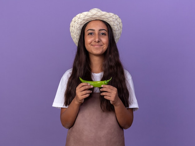 Jeune fille de jardinier en tablier et chapeau d'été tenant le piment vert souriant avec un visage heureux debout sur un mur violet