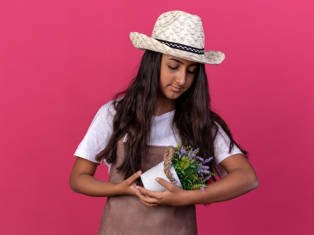 Jeune Fille De Jardinier En Tablier Et Chapeau D'été Tenant Une Plante En Pot En Regardant Avec Amour Souriant Debout Sur Un Mur Rose