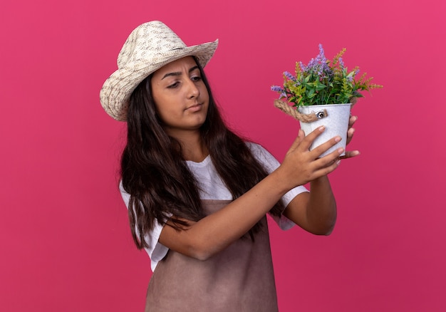 Jeune fille de jardinier en tablier et chapeau d'été tenant une plante en pot à la regarder intriguée debout sur un mur rose