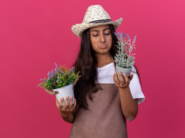 Jeune fille de jardinier en tablier et chapeau d'été tenant des plantes en pot en le regardant être mécontent et triste debout sur un mur rose