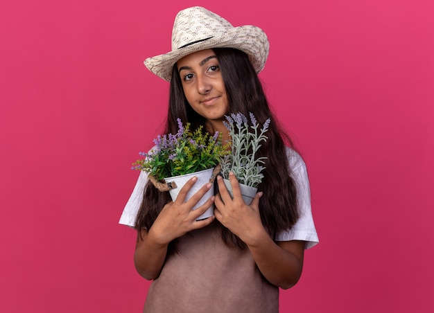 Jeune fille de jardinier en tablier et chapeau d'été tenant des plantes en pot souriant avec un visage heureux debout sur un mur rose