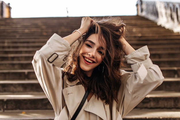 Jeune fille joyeuse avec une coiffure ondulée foncée et des lèvres rouges, portant un trench beige, souriante, décoiffant les cheveux et regardant à l'avant contre le mur des escaliers de la vieille ville