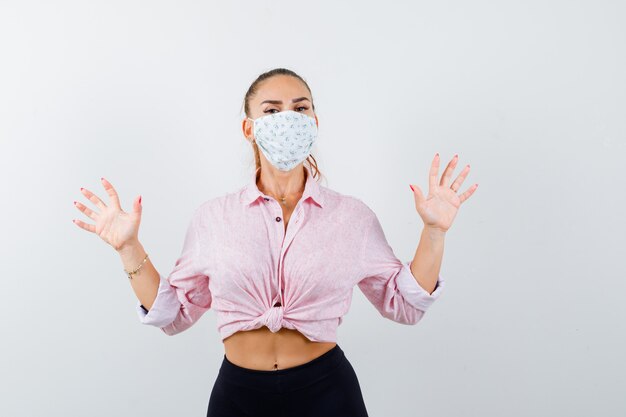 Jeune fille levant les paumes en geste de capitulation en chemisier rose, pantalon noir, masque et air effrayé, vue de face.